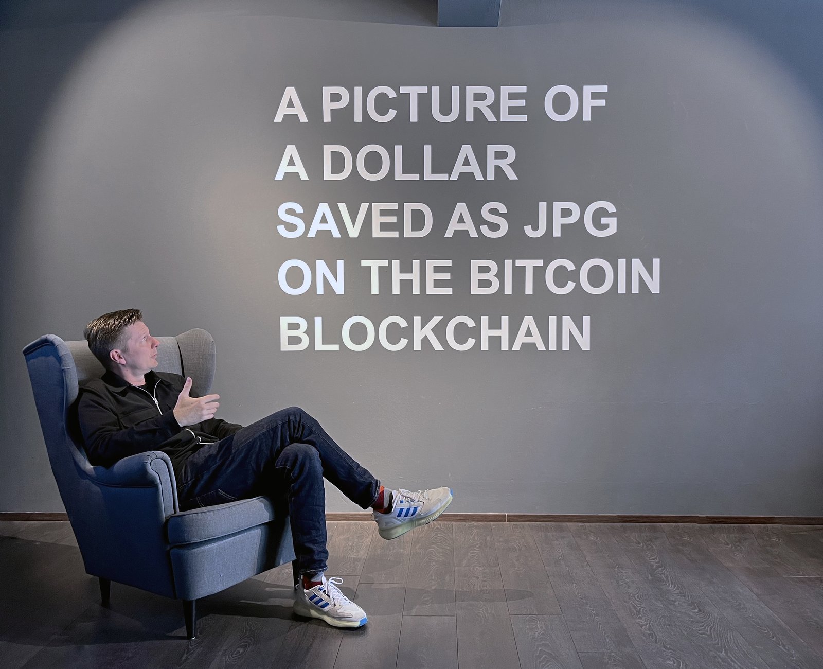 TRIBUTE TO GUY — the artist seated before the wall text A PICTURE OF A DOLLAR SAVED AS JPG ON THE BITCOIN BLOCKCHAIN